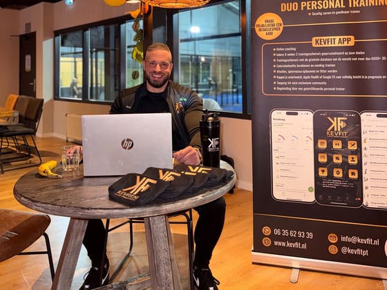 Kevin achter zijn laptop aan tafel, met de KEVFIT-banner ernaast